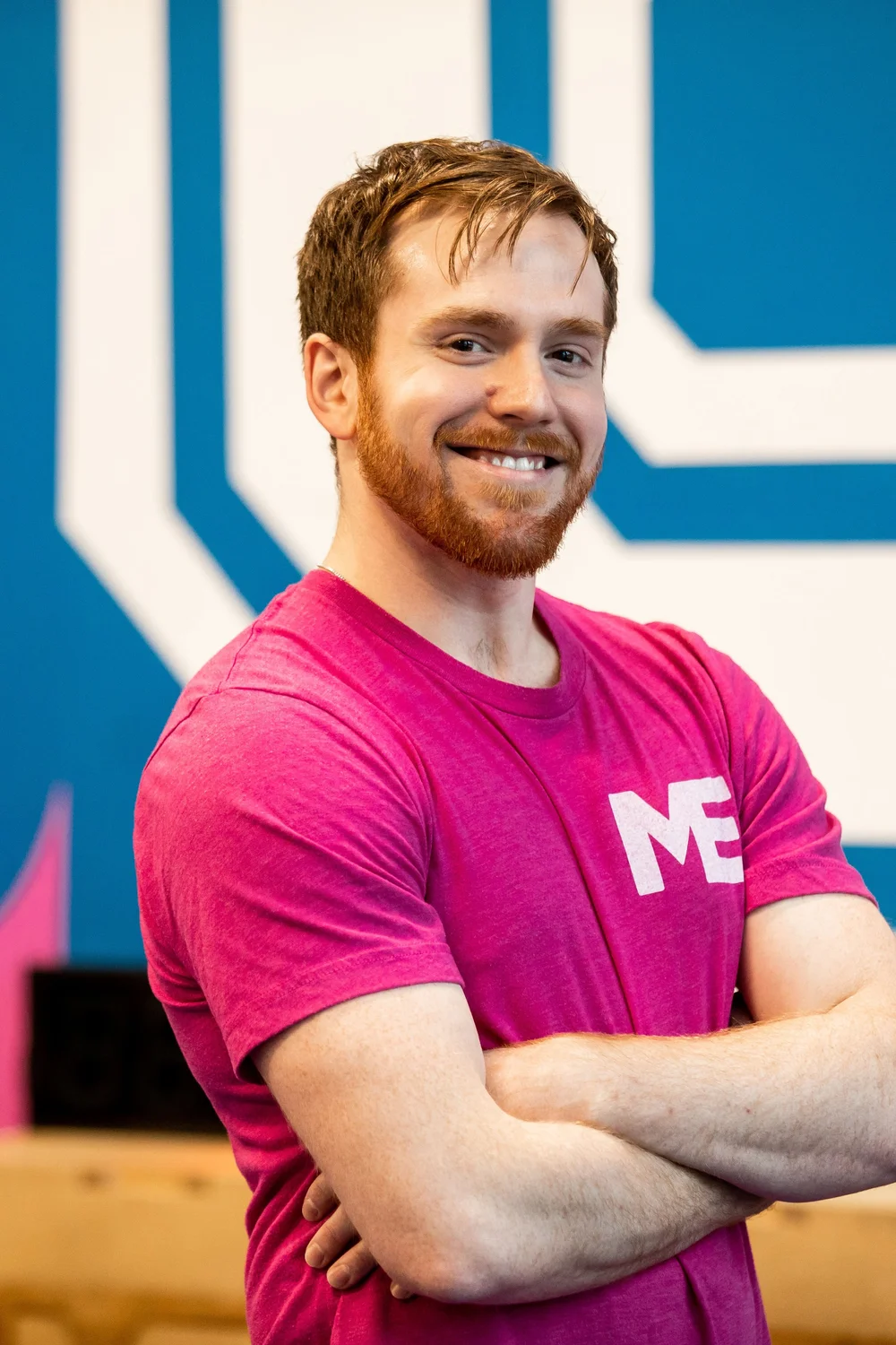 Billy, fitness coach at Studio ME Fitness, smiling with arms crossed in front of the Studio ME mural in Northeast Minneapolis.