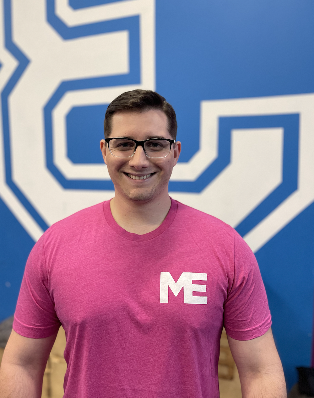 Luke, strength coach at Studio ME Fitness, smiling in a black Studio ME shirt in front of the pink and blue studio mural in Northeast Minneapolis.