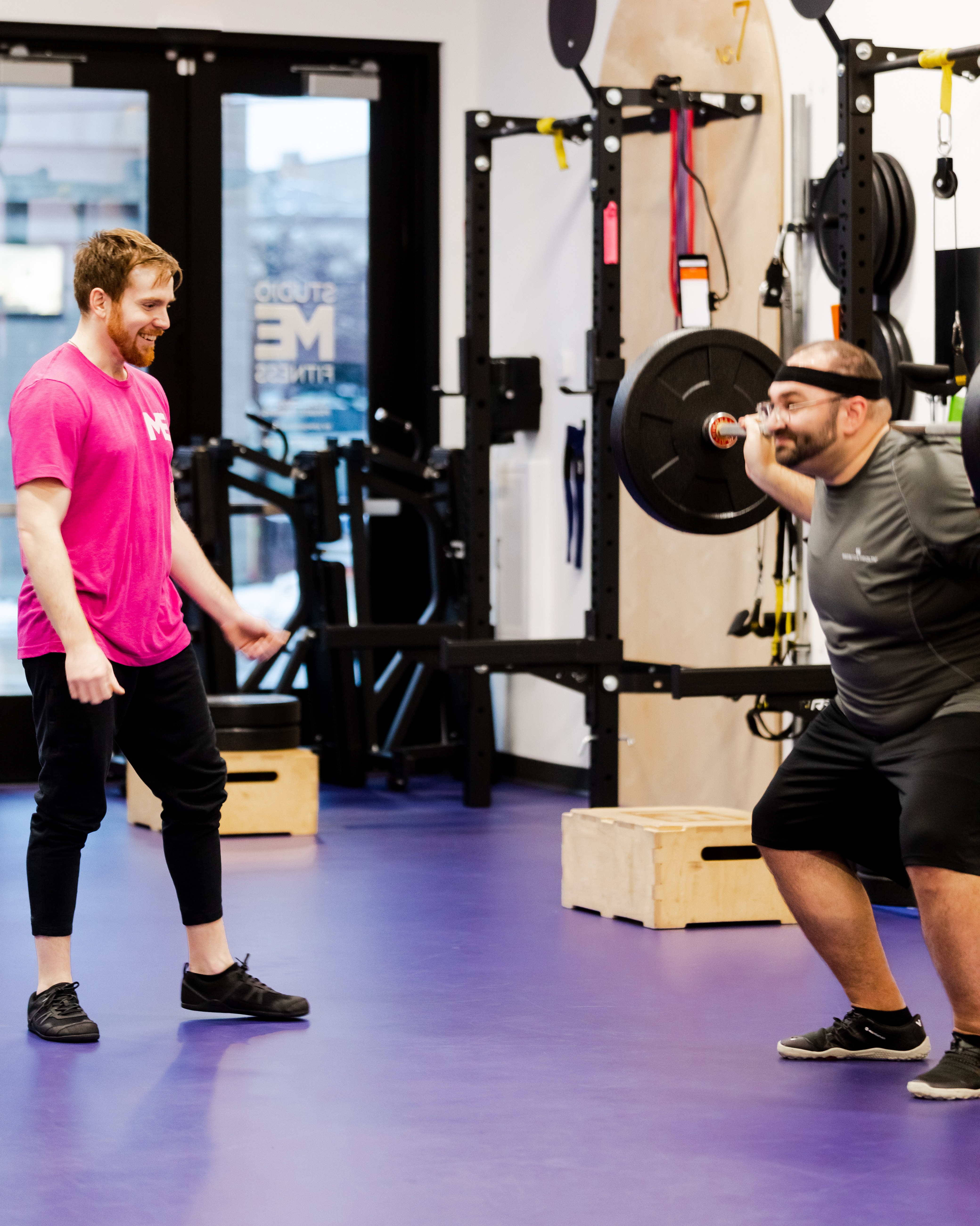 Studio ME Fitness coach in a pink shirt guiding a client through a barbell squat on the purple gym floor at our Northeast Minneapolis personal training studio.