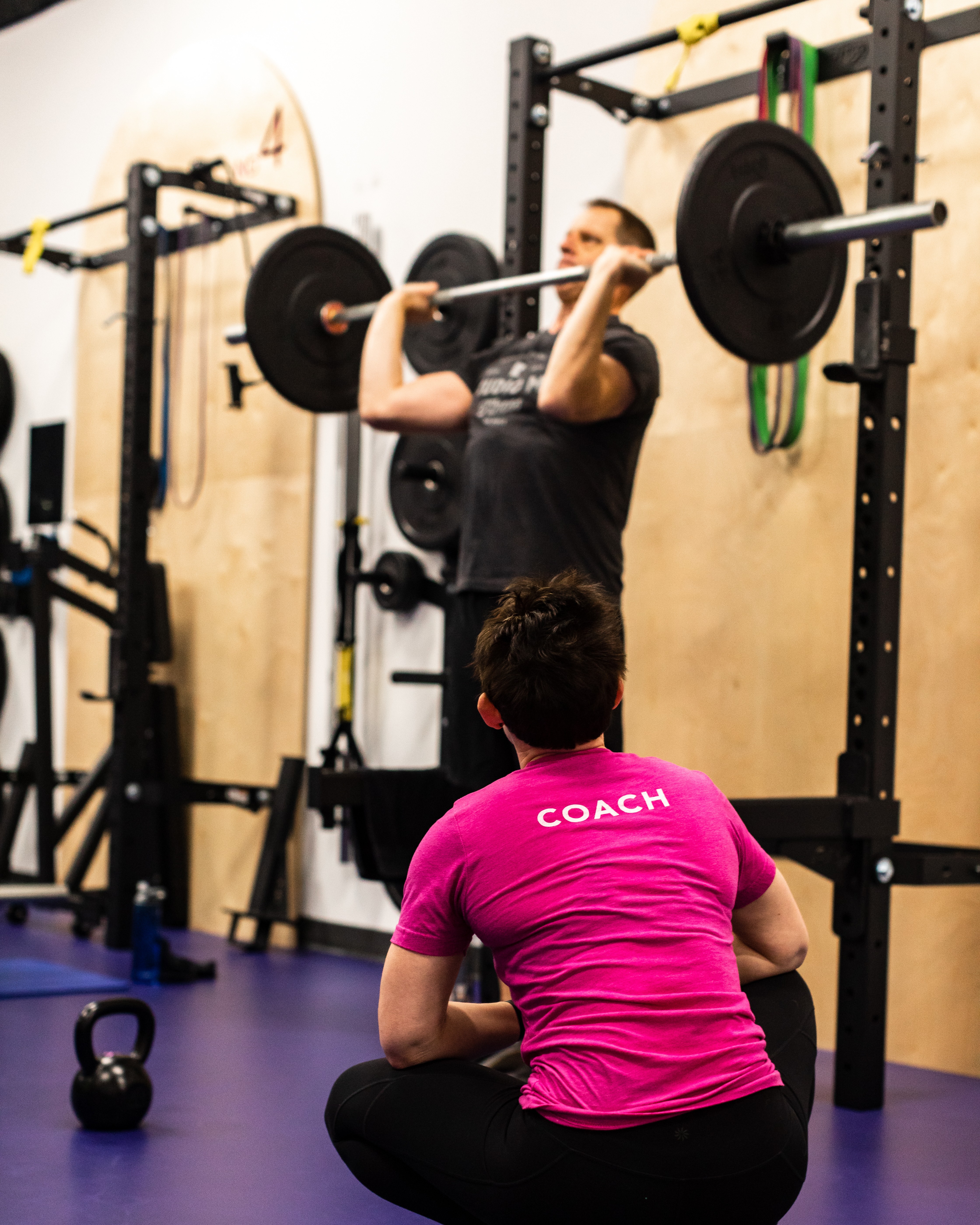 Studio ME Fitness coach in a pink COACH shirt observing a client performing a barbell back squat in the power rack during a semi-private training session in Northeast Minneapolis.