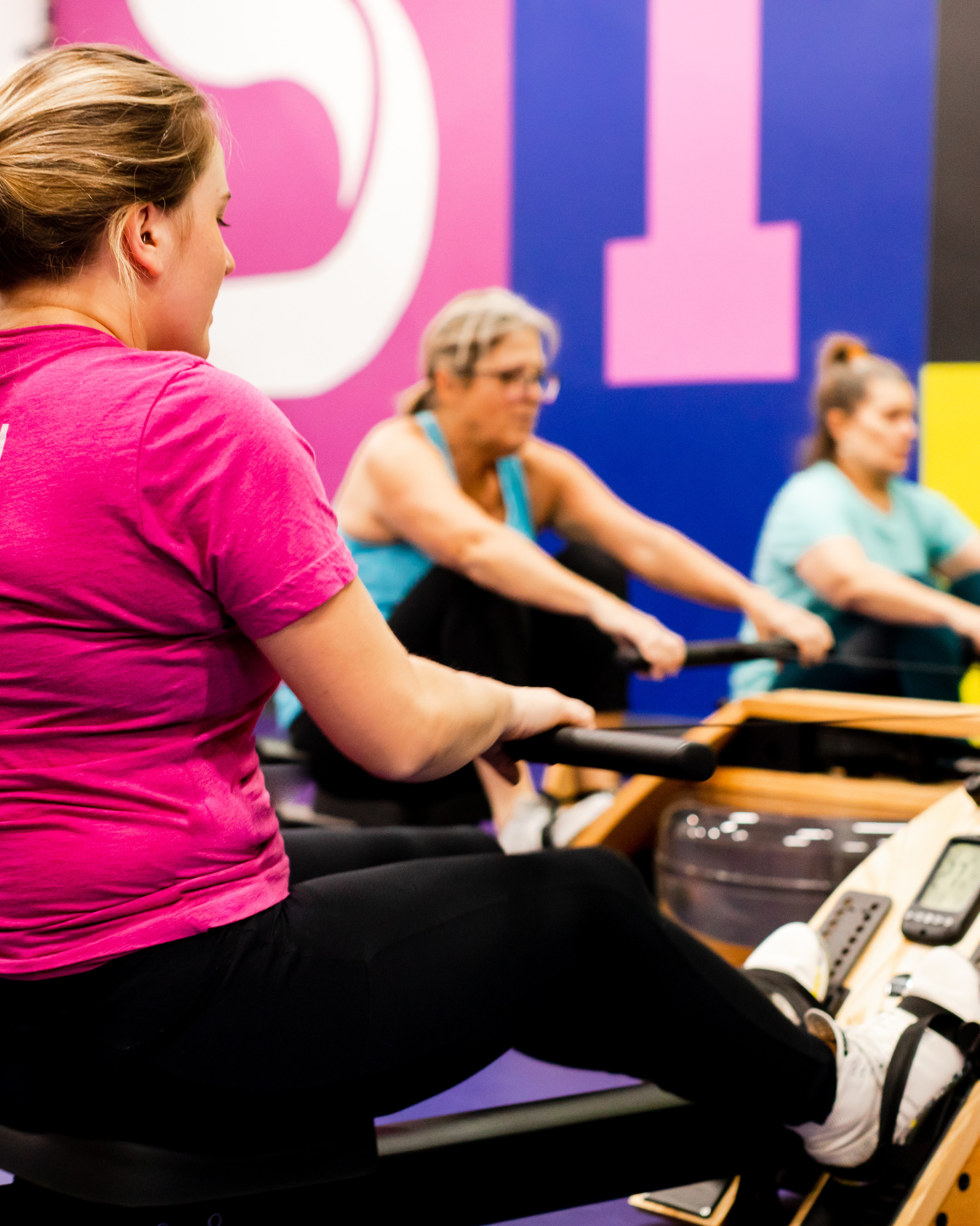 Three members rowing together during a small group training session at Studio ME Fitness in Northeast Minneapolis, with the studio's pink and purple branding on the wall behind them.