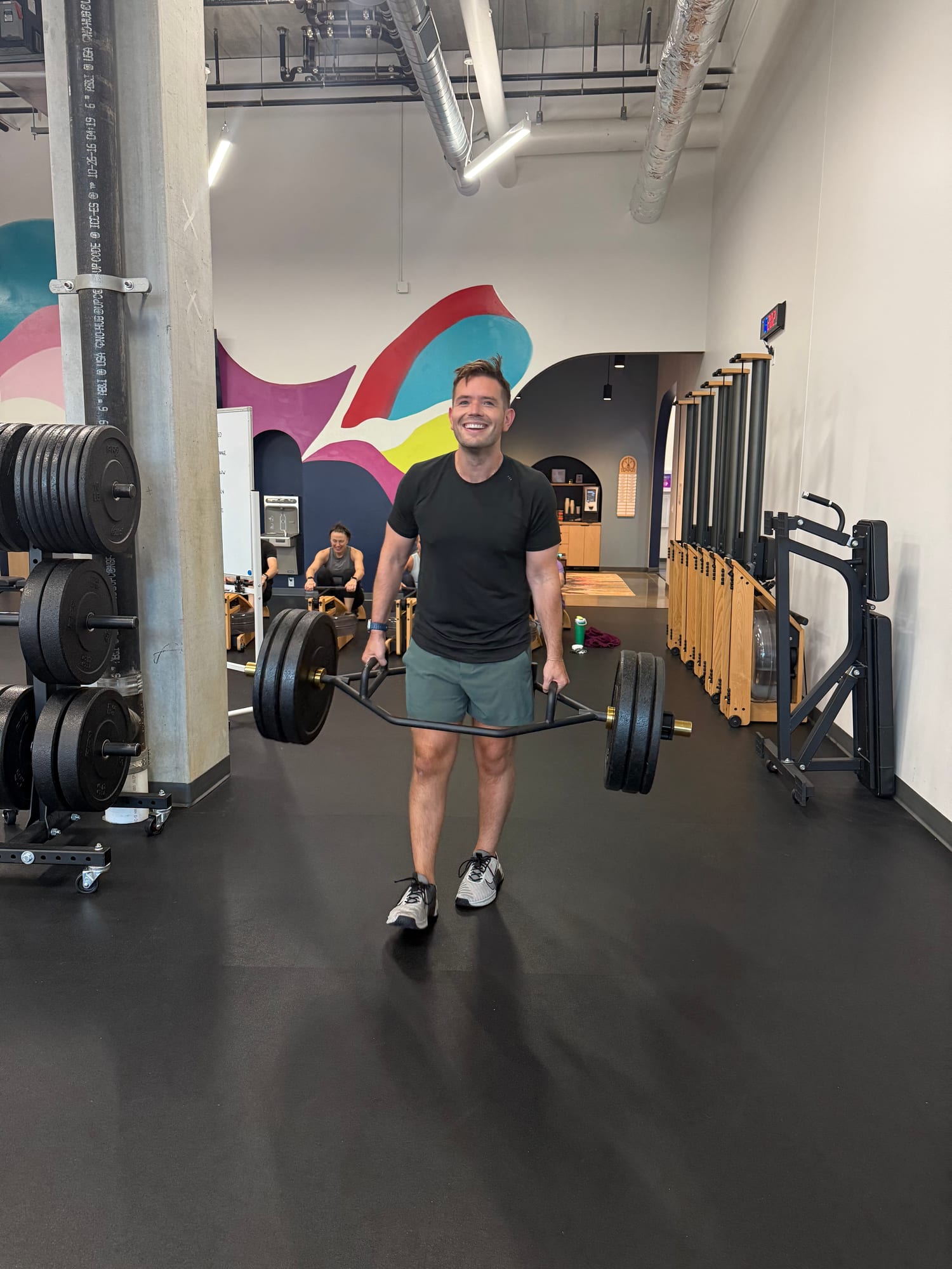"Man smiling while walking with a loaded trap bar during a farmer's carry exercise in a gym, with a rower and colorful mural in the background."