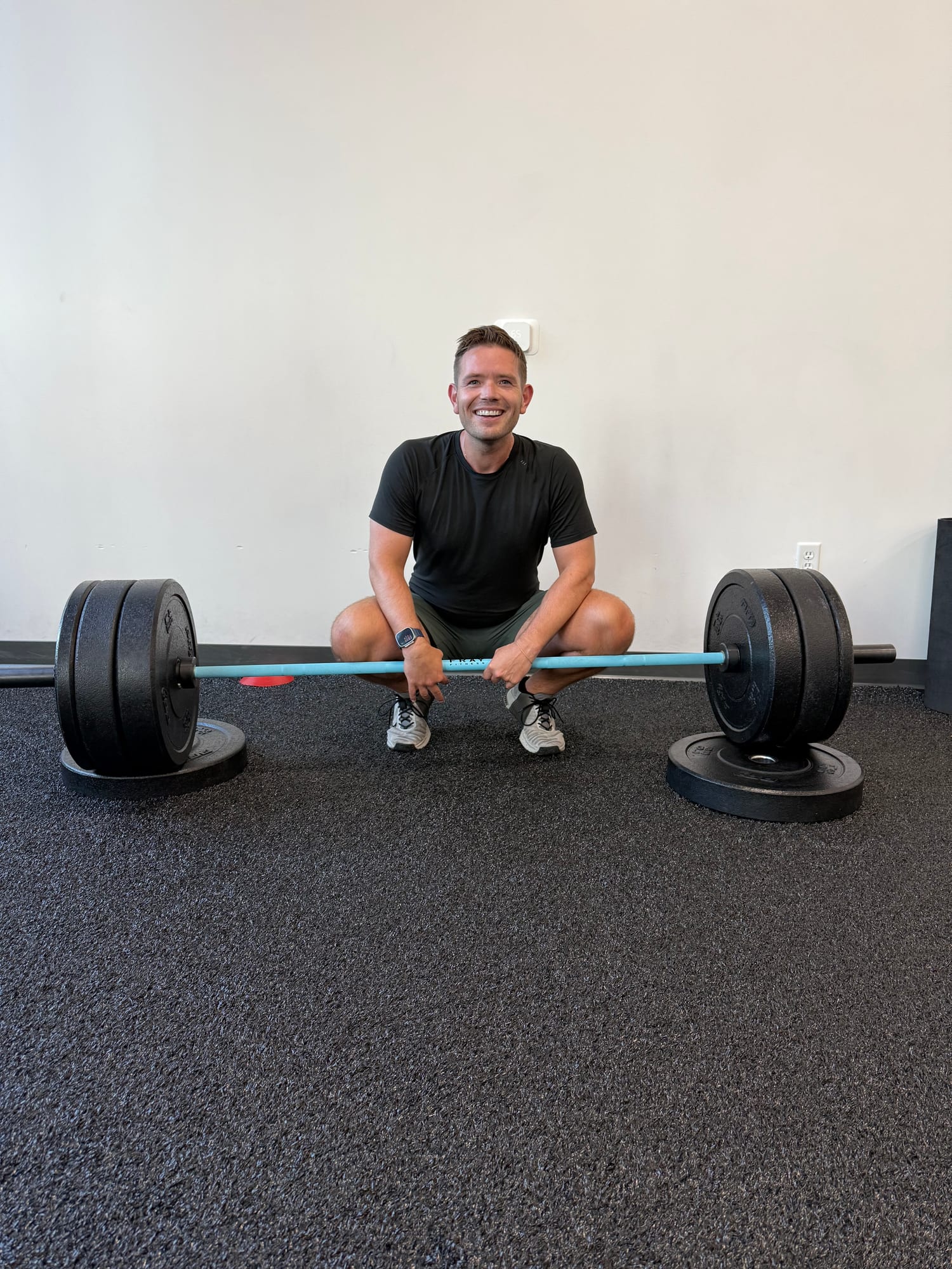 "Man in a squat position holding a barbell with weight plates, smiling at the camera in a gym setting."